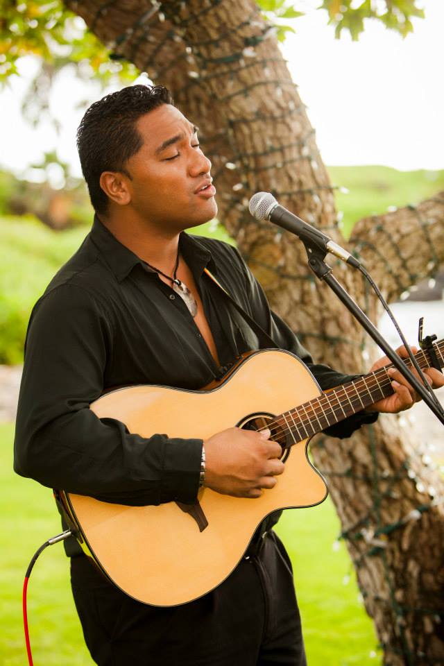 Kaniala Masoe singing into a microphone while playing acoustic guitar